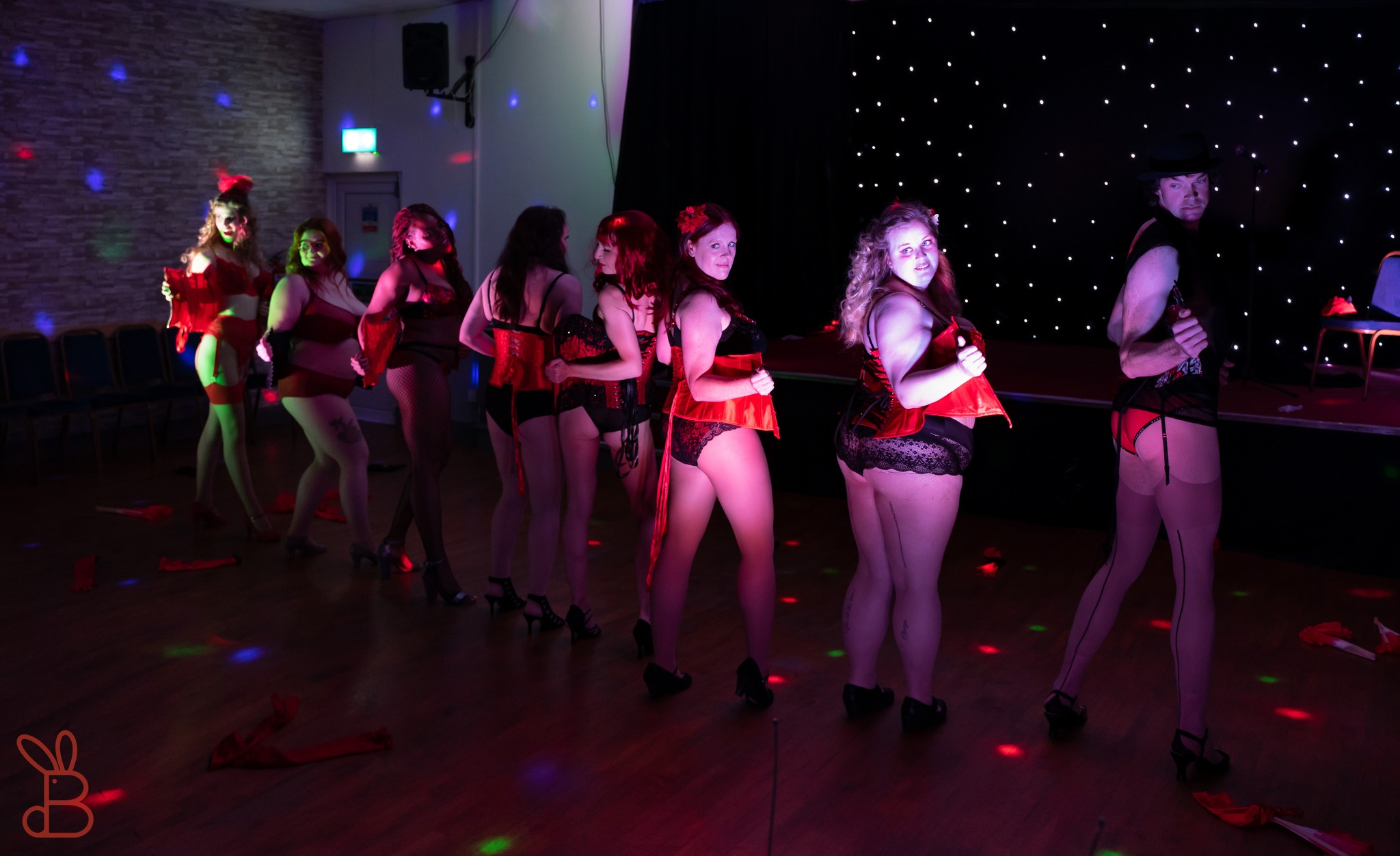 A row of eight dancers stand with their backs to the audience. They are a mix of body shapes and genders and are gazing over their shoulders at the audience while cheekily undoing their corsets.